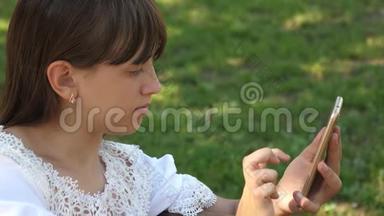 漂亮的女孩用智能手机在一个美丽的绿色公园的长凳上写了一封信。 慢动作。 少年女孩
